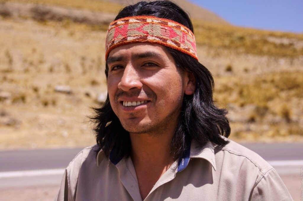 Portrait of a man wearing a traditional Andean headband, smiling with a mountainous landscape in the background.