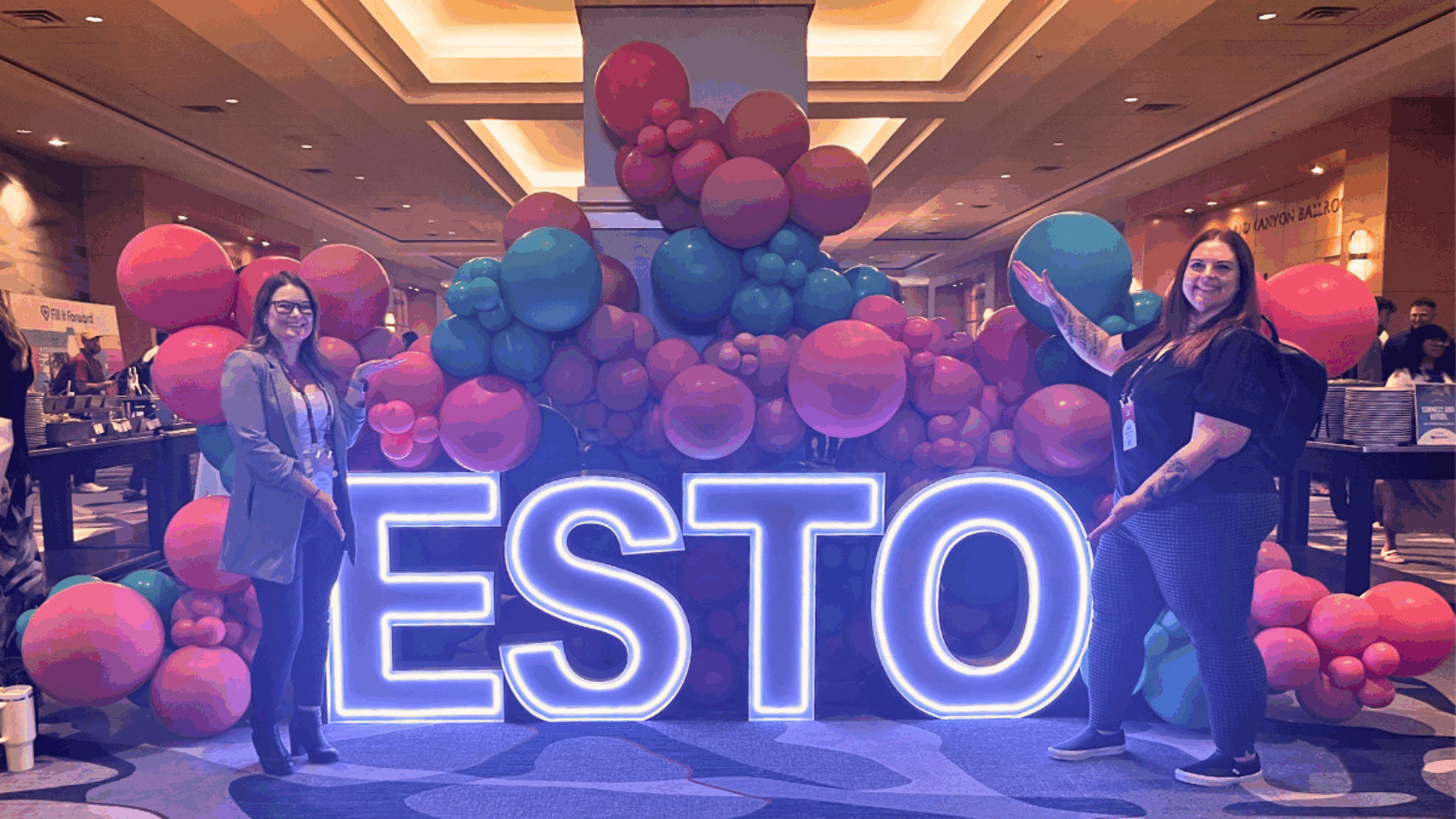 Two women pose beside large glowing “ESTO” letters in front of a vibrant balloon display at a conference event in a hotel lobby.