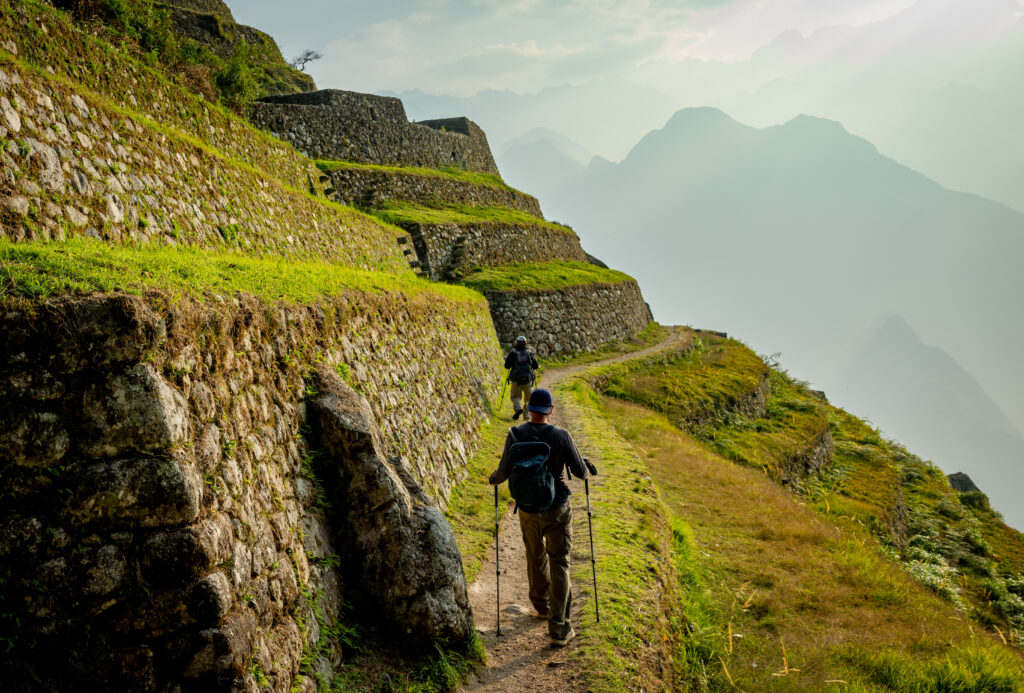 Two hikers walking along ancient stone terraces on the Inca Trail, surrounded by vibrant green grass and misty mountain views.