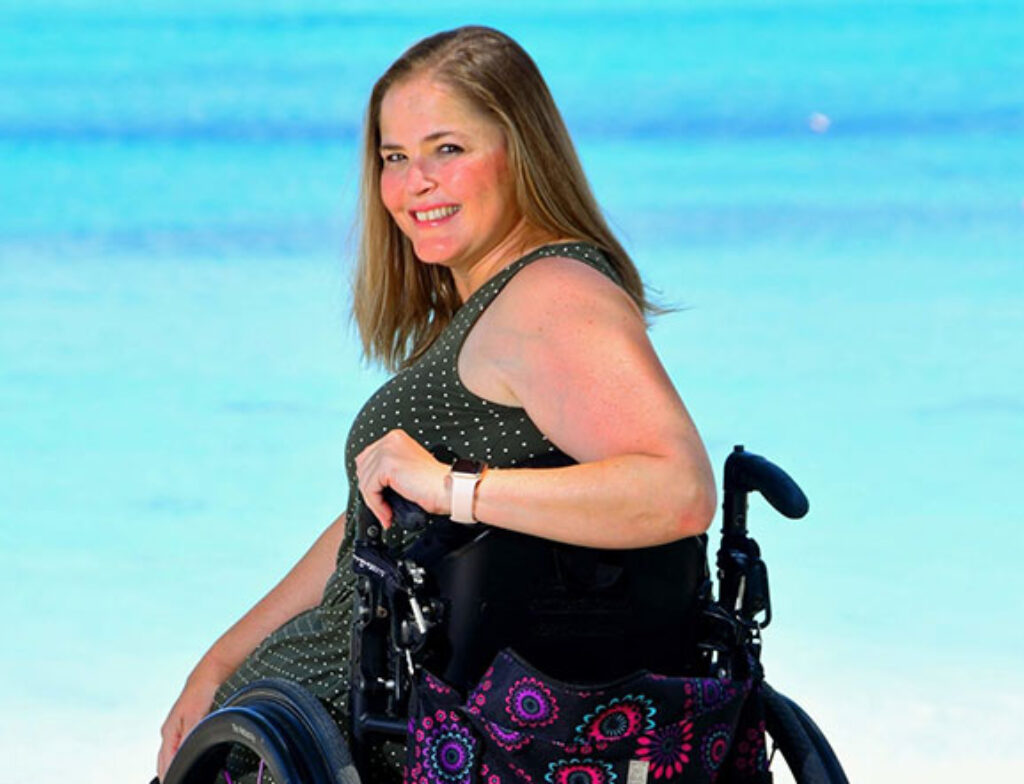 Kristy Durso smiling in a wheelchair by turquoise ocean water, wearing a sleeveless polka-dot dress.