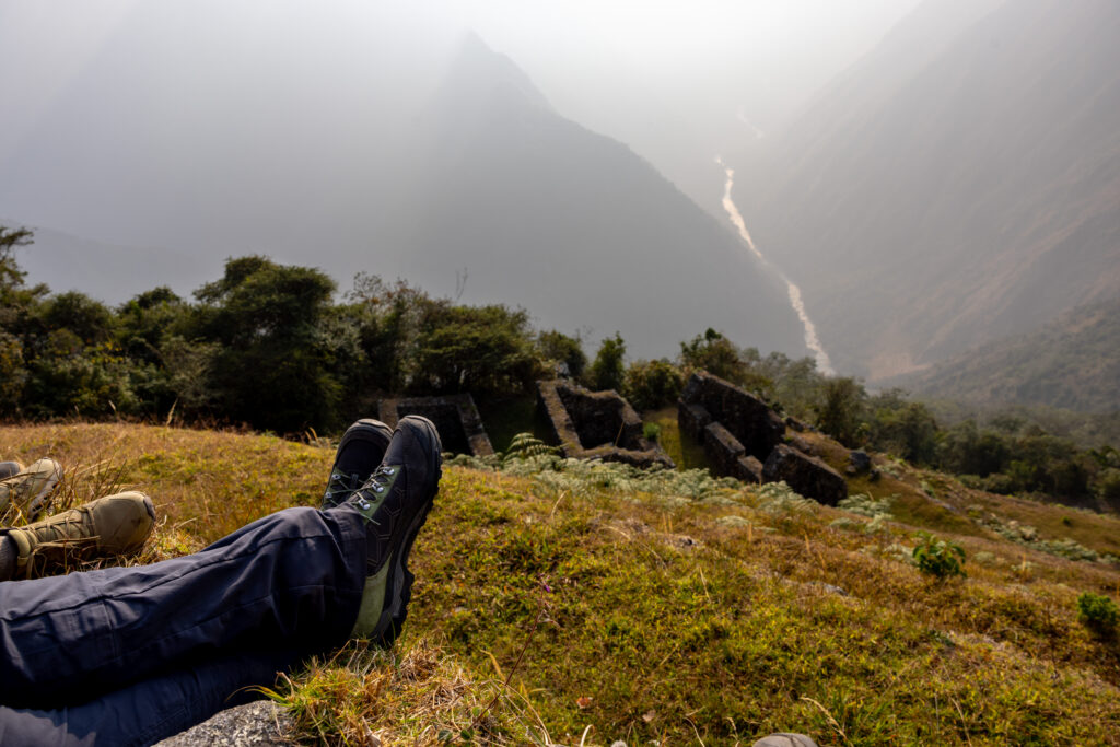 A pair of hikers resting on the Inca Trail, overlooking a scenic valley with ruins and a winding river below, symbolizing peaceful coexistence with nature.