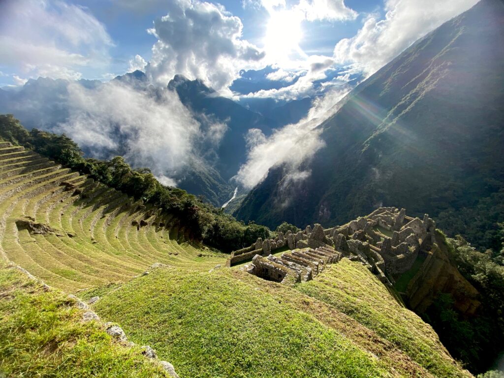 Terraced agricultural ruins on the Inca Trail with a misty mountain backdrop and sunlight breaking through the clouds.